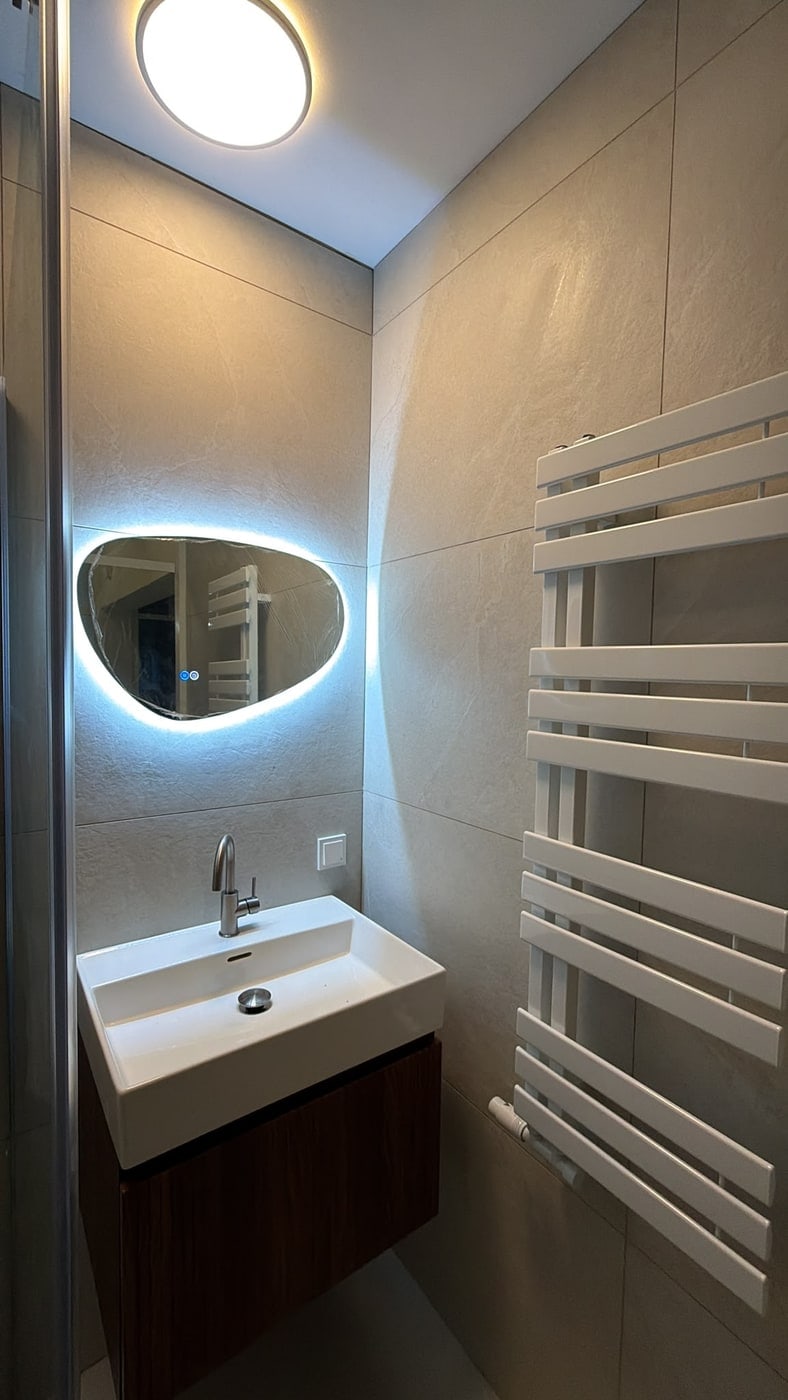 Afgewerkte badkamer met een wastafel op een houten onderkast, een verlichte spiegel en een witte handdoekradiator tegen grote lichte wandtegels.