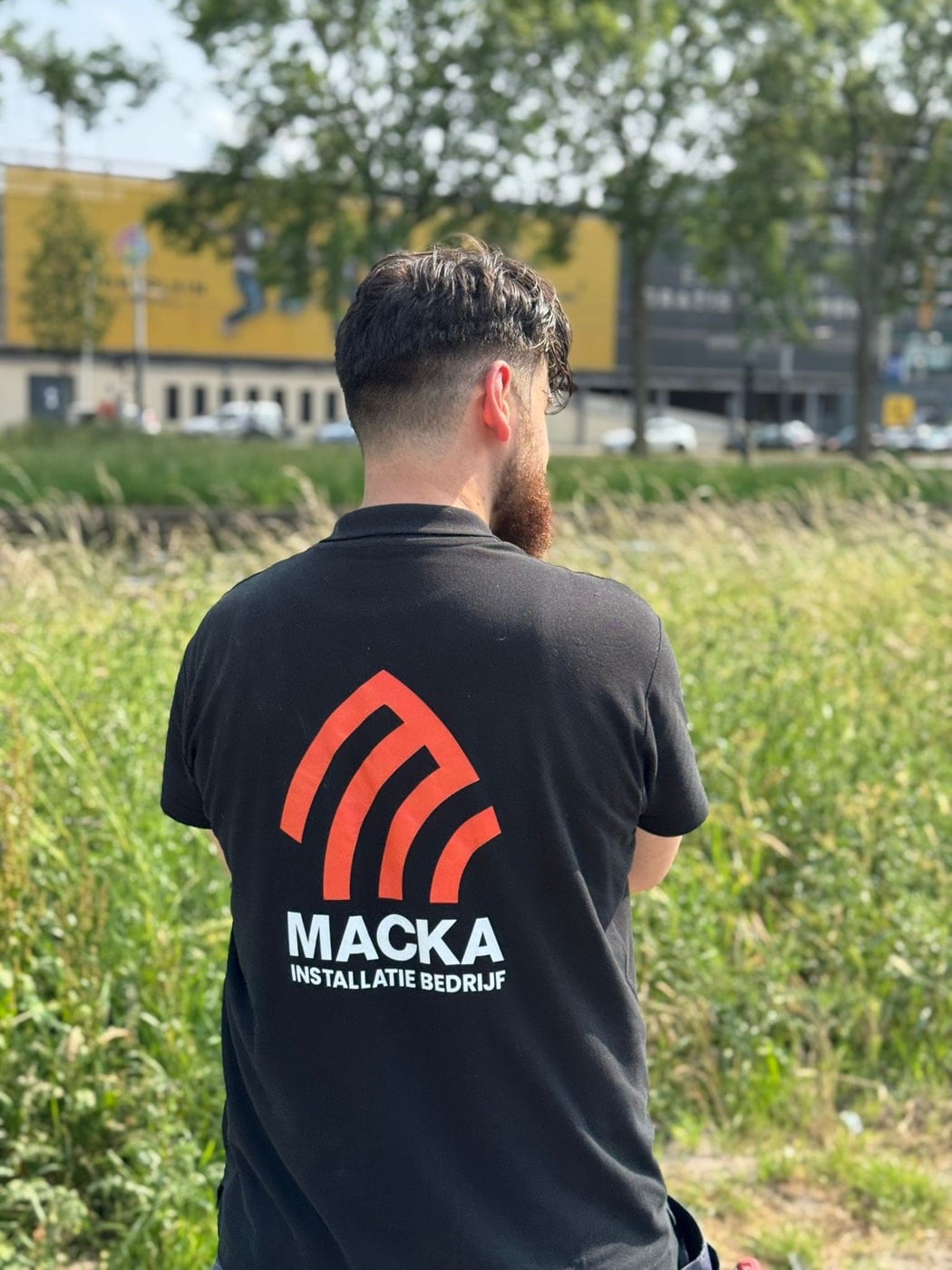Ömer van Macka Installatie van achteren gezien in bedrijfspolo met het logo op de rug: het oranje boogmerk boven de naam Macka Installatiebedrijf.