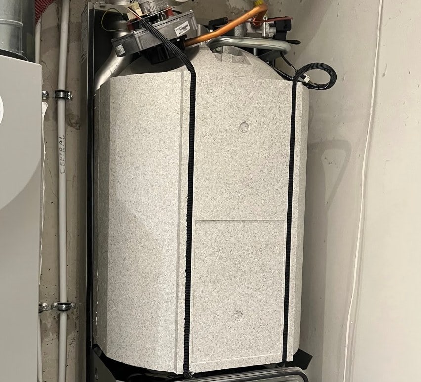 Cv-ketel in een opstelruimte met de mantel nog in transportfolie en spanbanden eromheen. Bovenop zijn de ventilator, het gasblok en de koperen gasaansluiting zichtbaar, met de rookgasafvoer links en de condensafvoer rechts.