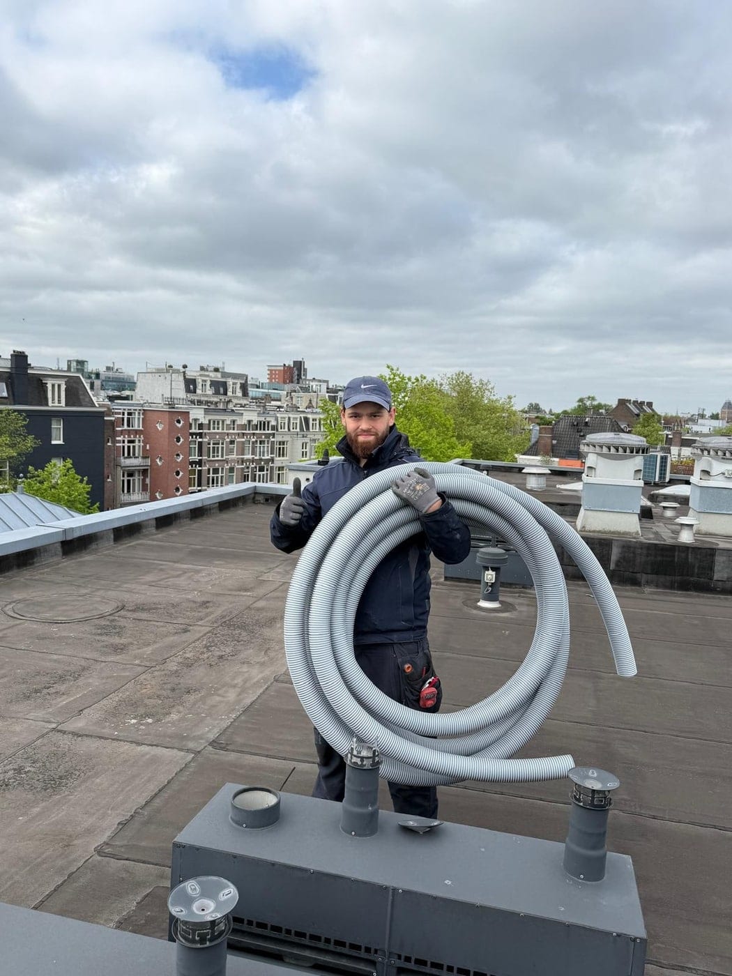 Ömer van Macka Installatie op een plat dak in Amsterdam met een rol flexibele ventilatieslang, met de daken van de stad op de achtergrond.