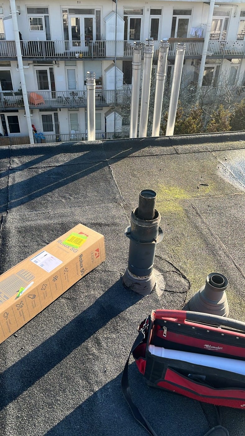 Rookgasafvoeren en dakdoorvoeren op een plat dak van een appartementencomplex, met gereedschapstas en een doos dakdoorvoermateriaal.