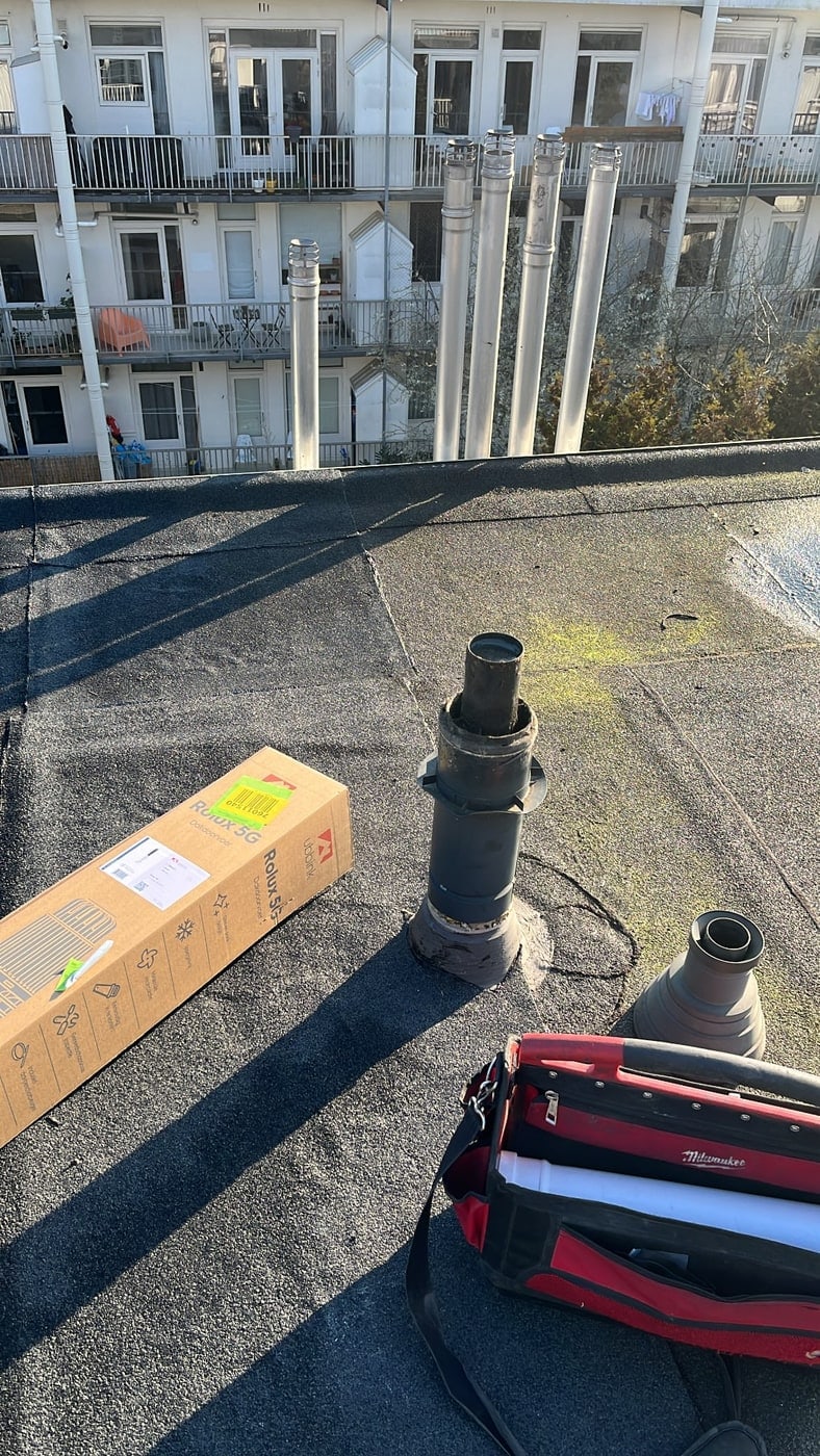 Rookgasafvoeren en dakdoorvoeren op een plat dak van een appartementencomplex, met gereedschapstas en een doos dakdoorvoermateriaal.