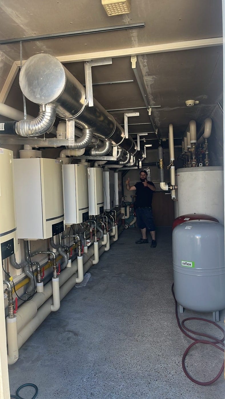 Volledige technische ruimte met acht cv-ketels in cascade langs de wand, met kanaalwerk langs het plafond en een expansievat.