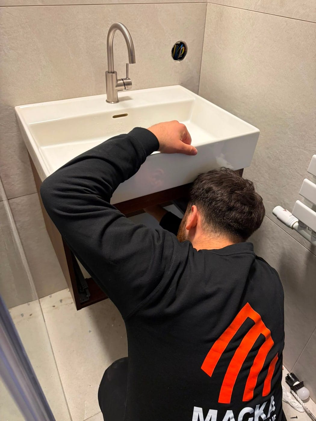 Ömer van Macka Installatie in bedrijfskleding met het oranje logo op de rug, bezig met de wateraansluiting onder een pas geplaatste wastafel.